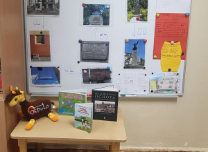 Tablica z pracami i zdjęciami związanymi z dzielnicą Ochota, ilustracjami i notatkami dzieci; na stoliku poniżej książki o Ochocie oraz maskotka żyrafy.