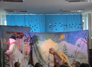 zimowa scena teatrzyku w przedszkolu: bałwanek i żyrafa na tle bajkowej, śnieżnej dekoracji z górami, miastem i gwiazdami, przed zgromadzonymi dziećmi.