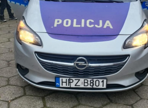 Radiowóz policyjny zaparkowany przed przedszkolem, z włączonymi niebieskimi światłami; wokół samochodu stoją dzieci oglądające pojazd podczas spotkania z policjantami.